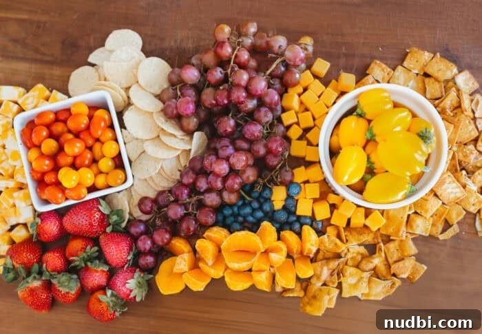Overhead view of a colorful snack board with Sunset® Sprinkles tomatoes, yellow grape tomatoes, strawberries, grapes, blueberries, cheese cubes, mandarin slices, crackers, and tortilla chips on a wooden board, perfect for kids.