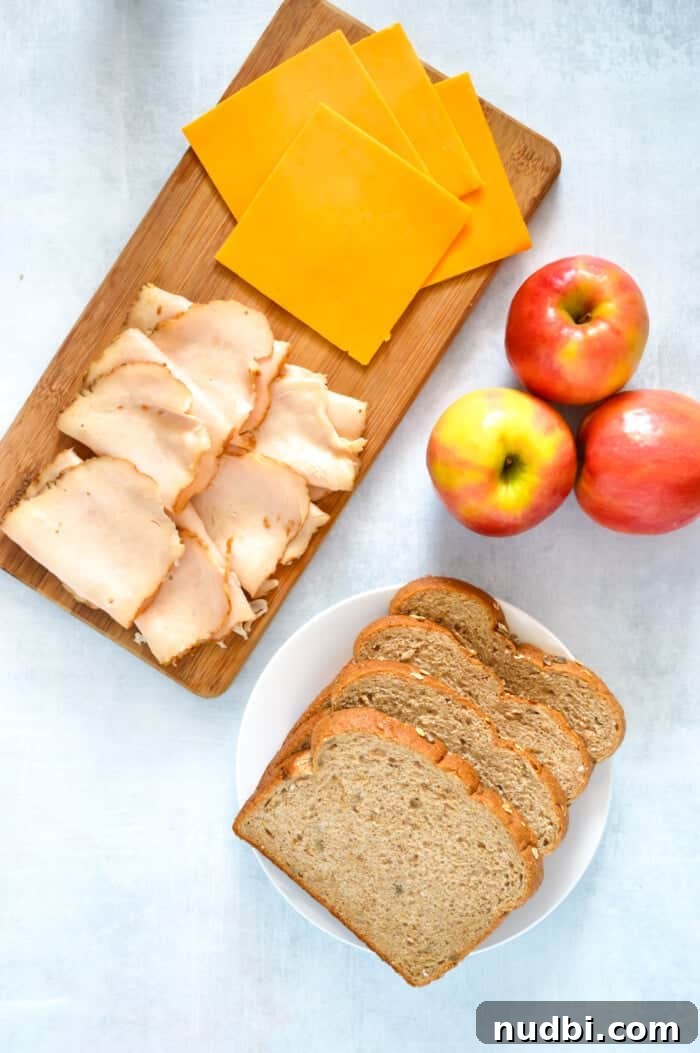 Assorted gourmet grilled cheese ingredients: slices of ham, cheddar cheese, fresh apple, and rustic bread.