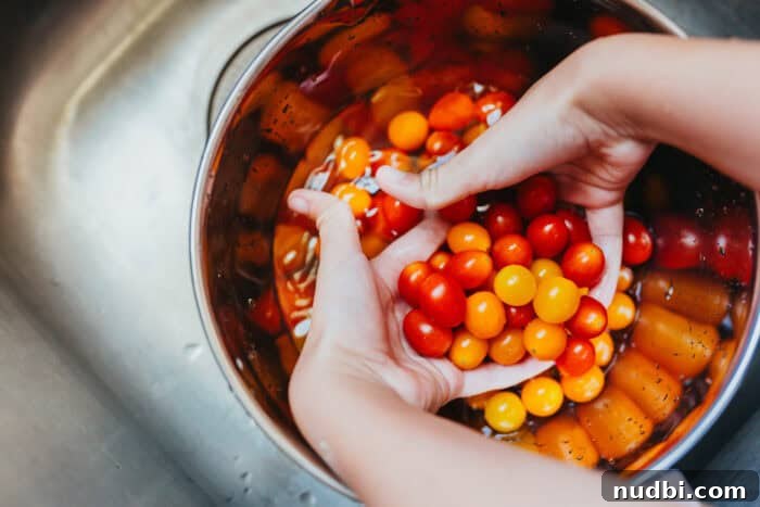 Close-up of small hands gently rinsing vibrant Sunset® Sprinkles teeny tiny tomatoes in a large clear bowl filled with water, positioned over a modern kitchen sink. The act emphasizes fresh produce preparation.
