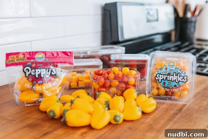 Assorted clear containers filled with vibrant back-to-school fridge staples: Sunset® Sprinkles tomatoes, Peppy's teeny tiny peppers, juicy grapes, ripe strawberries, and savory cheese cubes, all neatly arranged on a kitchen counter adjacent to a stove. The image highlights fresh and ready-to-eat snack components.