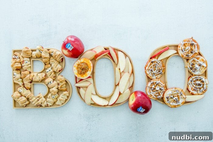 Halloween board spelling BOO with apple slices, cinnamon rolls shaped like mummies, and other festive treats.