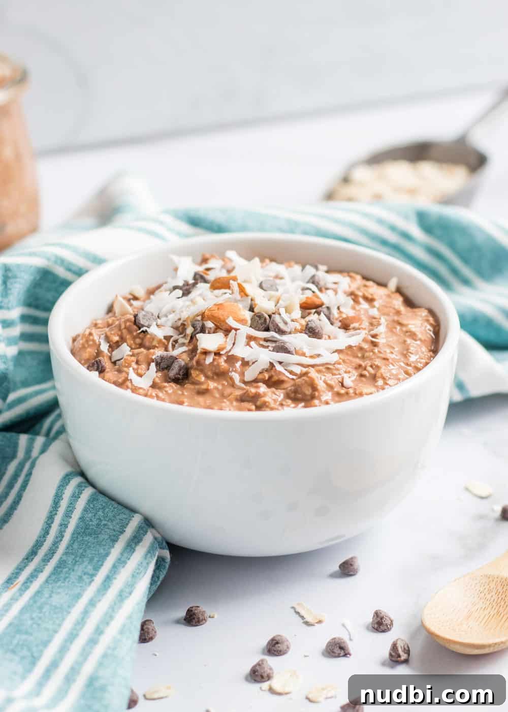 Almond Joy Overnight Oats Recipe — Chocolate, Coconut & Almonds 4 Almond joy overnight oats in white bowl with chocolate chips, coconut and almonds on top and spoon beside the bowl.