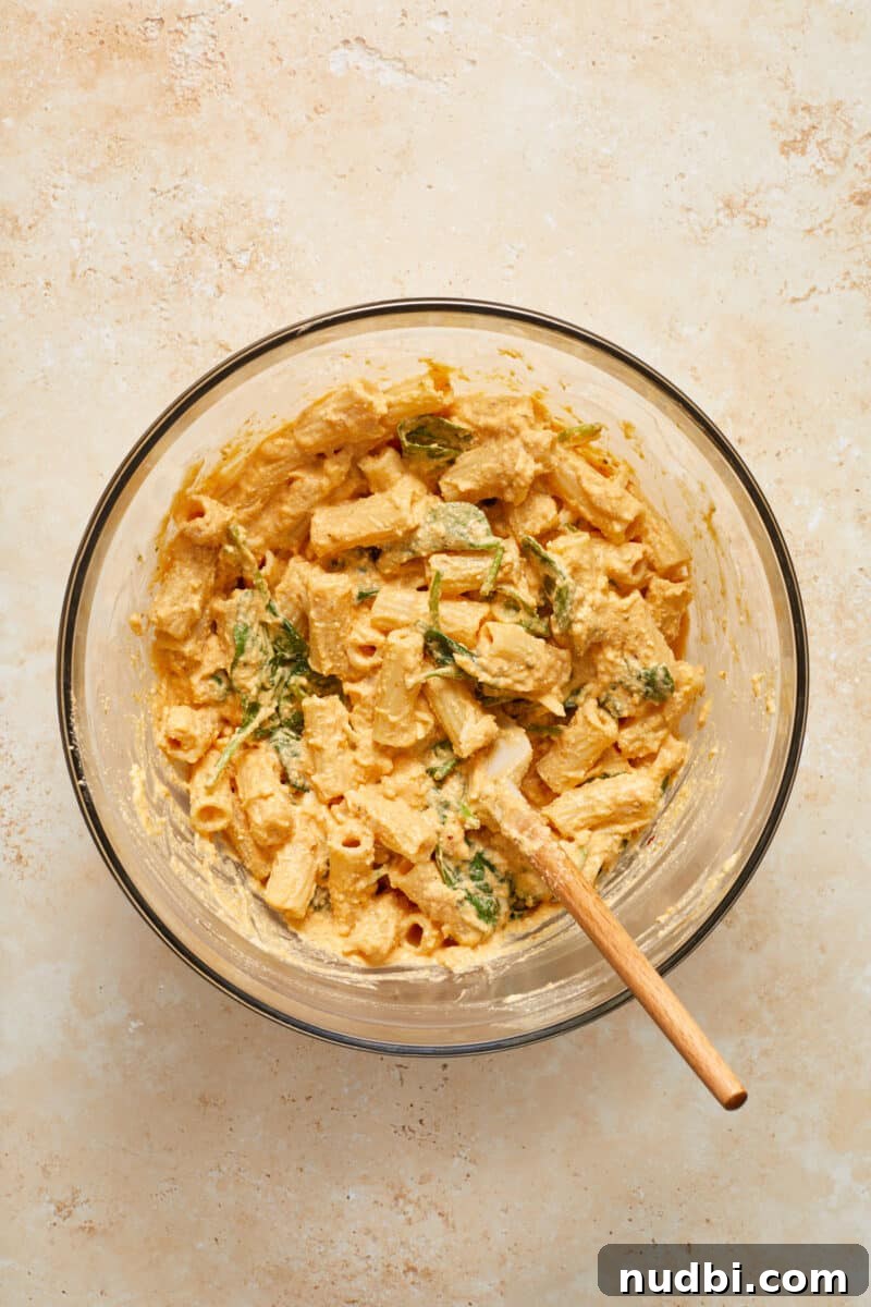 Creamy Pumpkin Ricotta Pasta Bake with Parmesan Crisp Topping 7 Pumpkin mixture tossed with pasta and spinach in glass mixing bowl with spatula.
