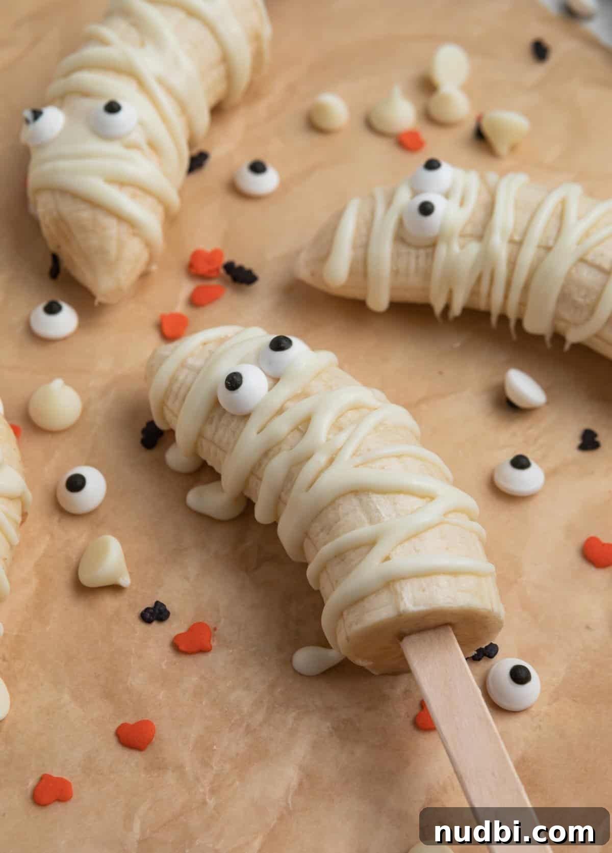 Spooky Banana Mummy Pops: Halloween Treats for Kids 3 Mummy banana popsicle on parchment with white chocolate chips and Halloween sprinkles surrounding.