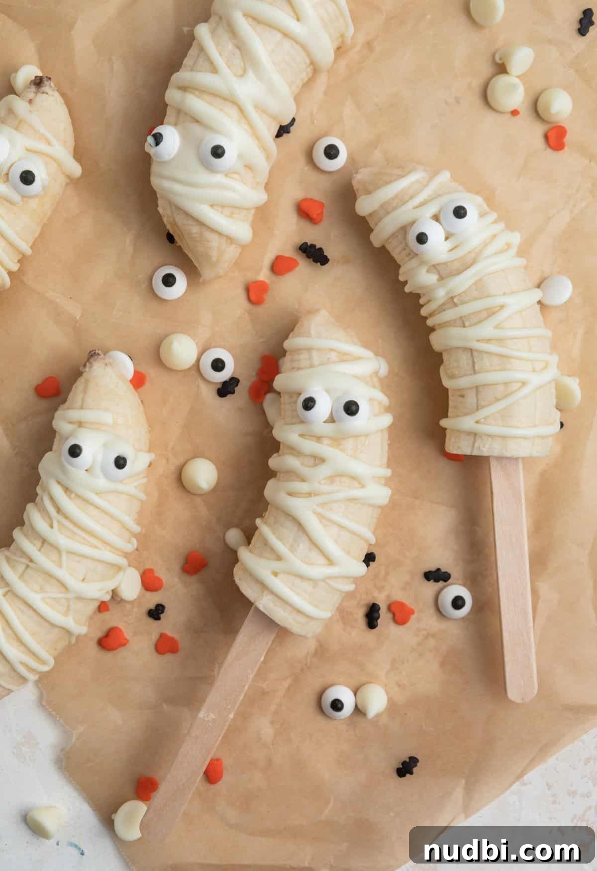 Spooky Banana Mummy Pops: Halloween Treats for Kids 4 Halloween banana mummies on brown parchment with Halloween sprinkles surrounding.