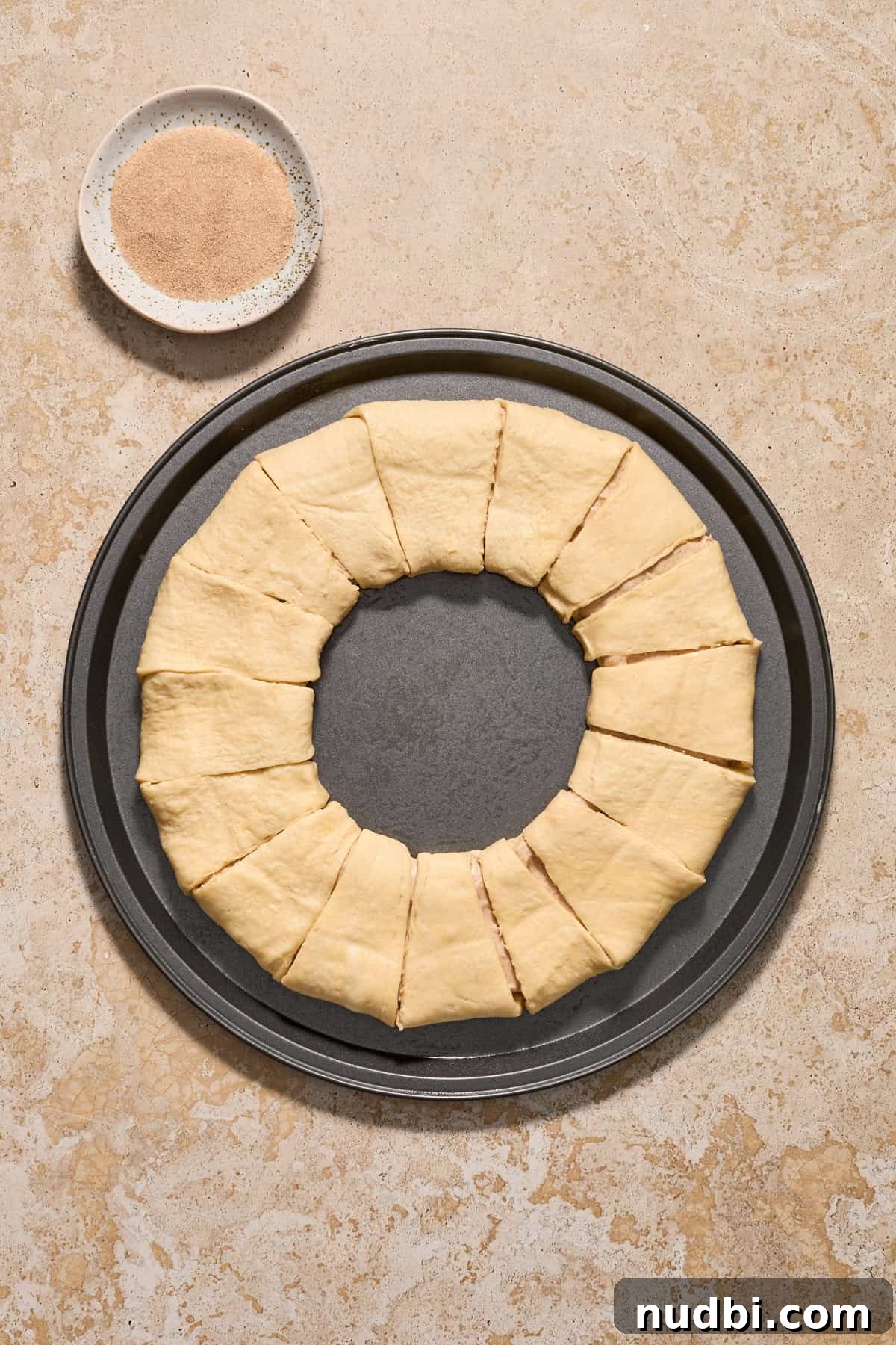 Cinnamon Crescent Roll Wreath Recipe — Festive Holiday Pastry 10 Crescent roll wreath on cookie sheet before baking with cinnamon and sugar in bowl beside it.