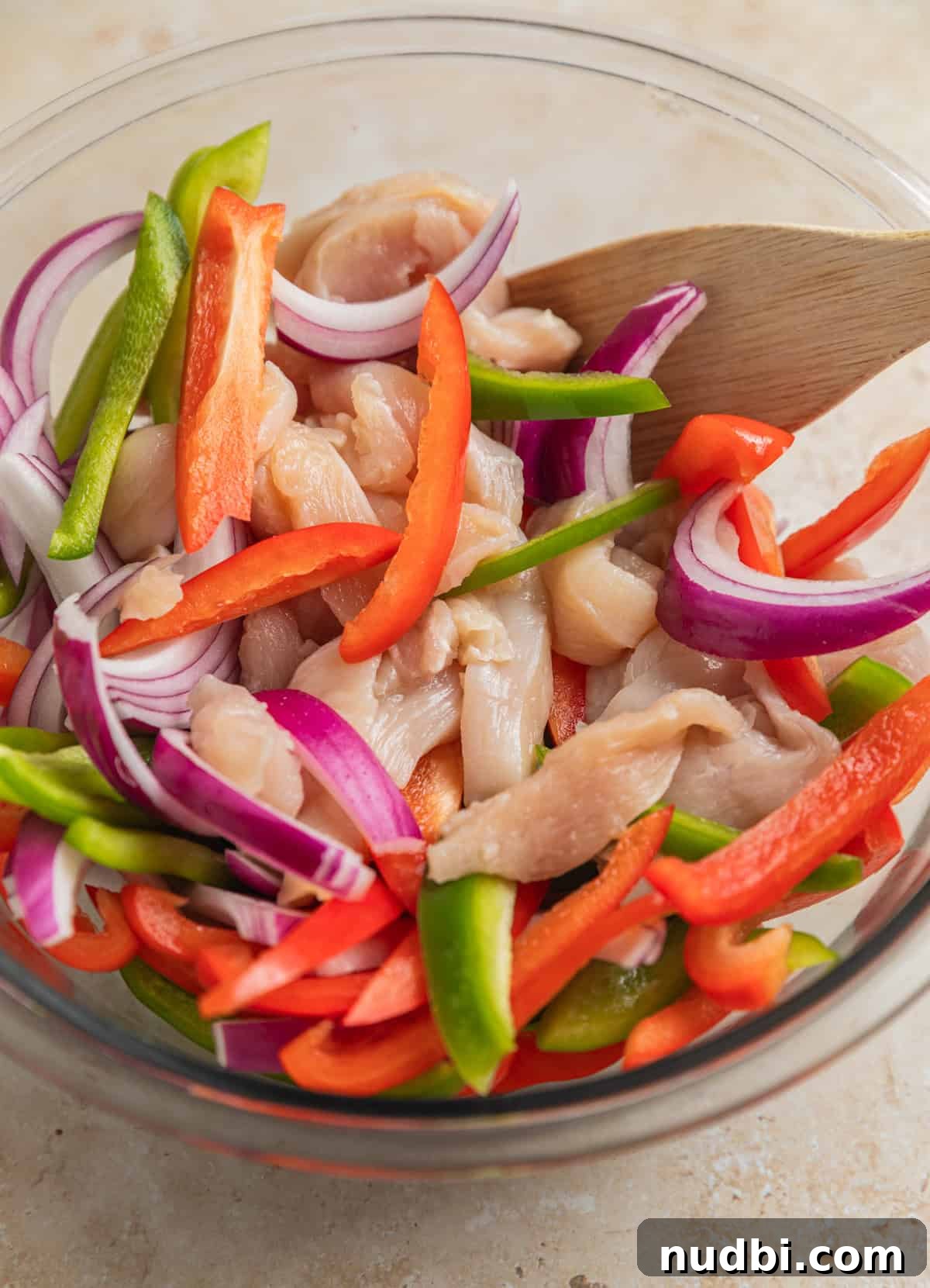 Honey-Lime Baked Chicken Fajitas Recipe for Flavorful Weeknights 4 Raw sliced chicken, sliced red and green bell peppers and red onion in glass bowl with wood spoon.