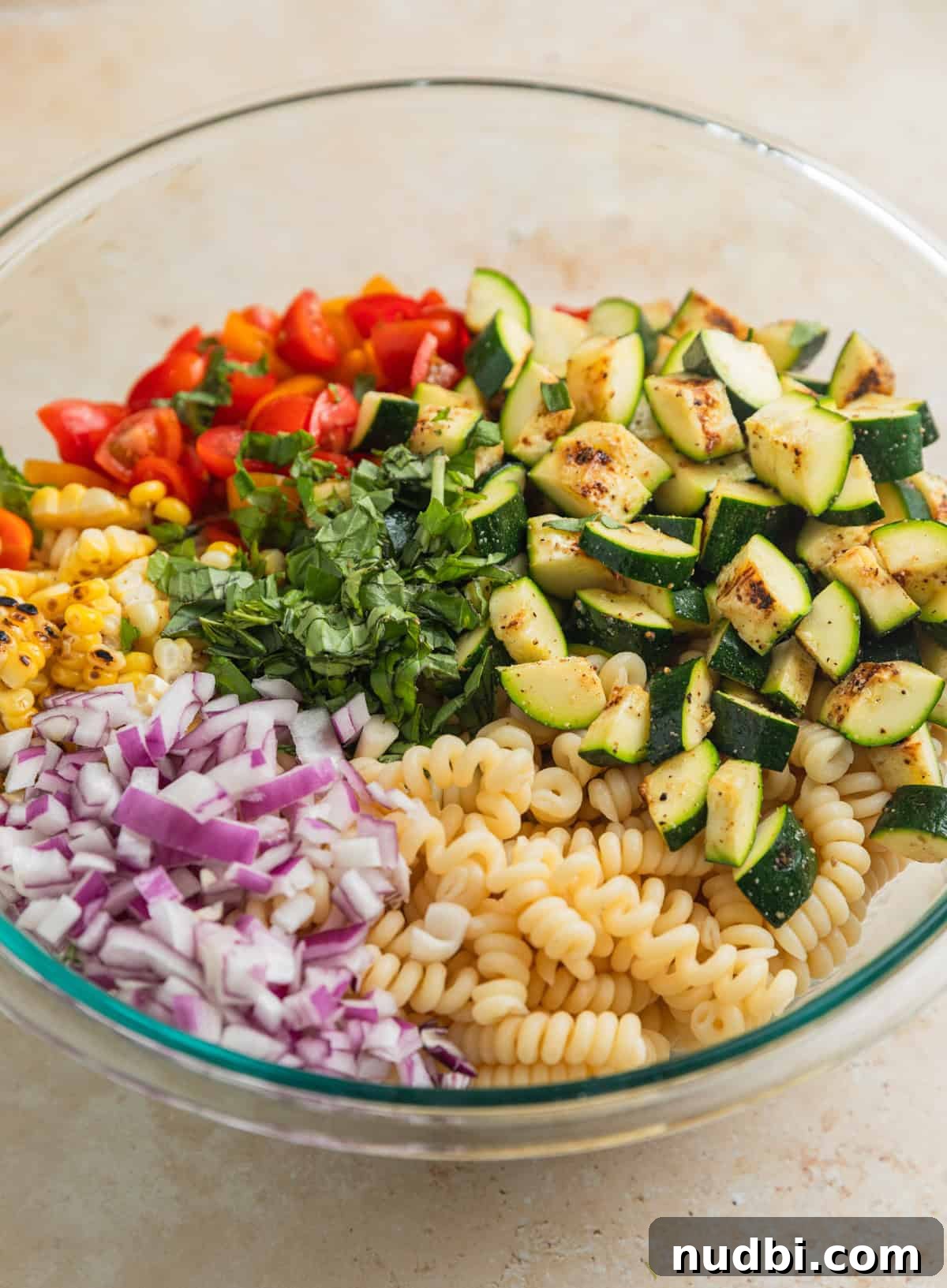 Grilled Zucchini Pasta Salad with Lemon Garlic Dressing 7 Cooked pasta, red onion, tomatoes, corn and zucchini in glass mixing bowl.