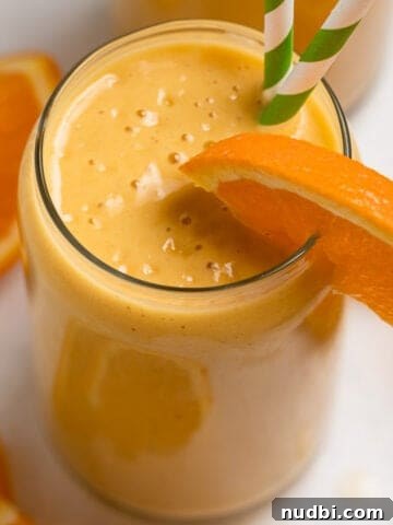 Orange banana smoothie in glass with orange slice to garnish and green striped straws.