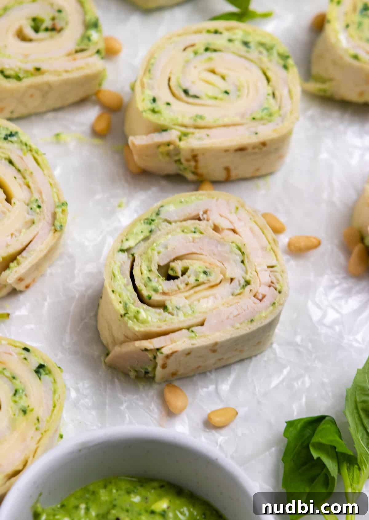 Smoked Provolone Turkey Pesto Pinwheels рецепт 5 Turkey pinwheels with pesto on wax paper with basil and pine nuts surrounding.