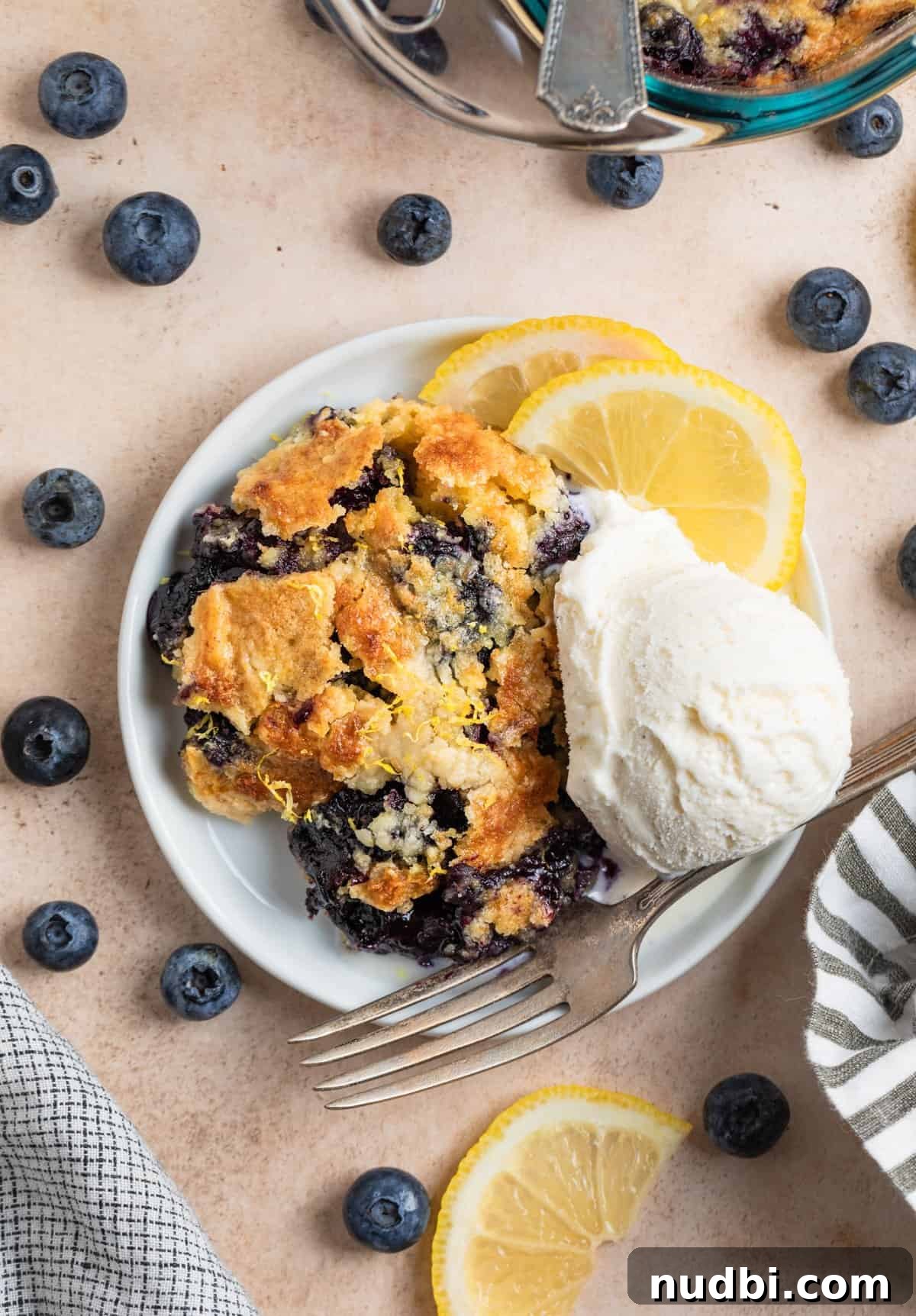 Lemon Blueberry Dump Cake Recipe for Quick Summer Desserts 3 Overhead view of piece of blueberry cobbler on white plate with ice cream and fresh blueberries surrounding.