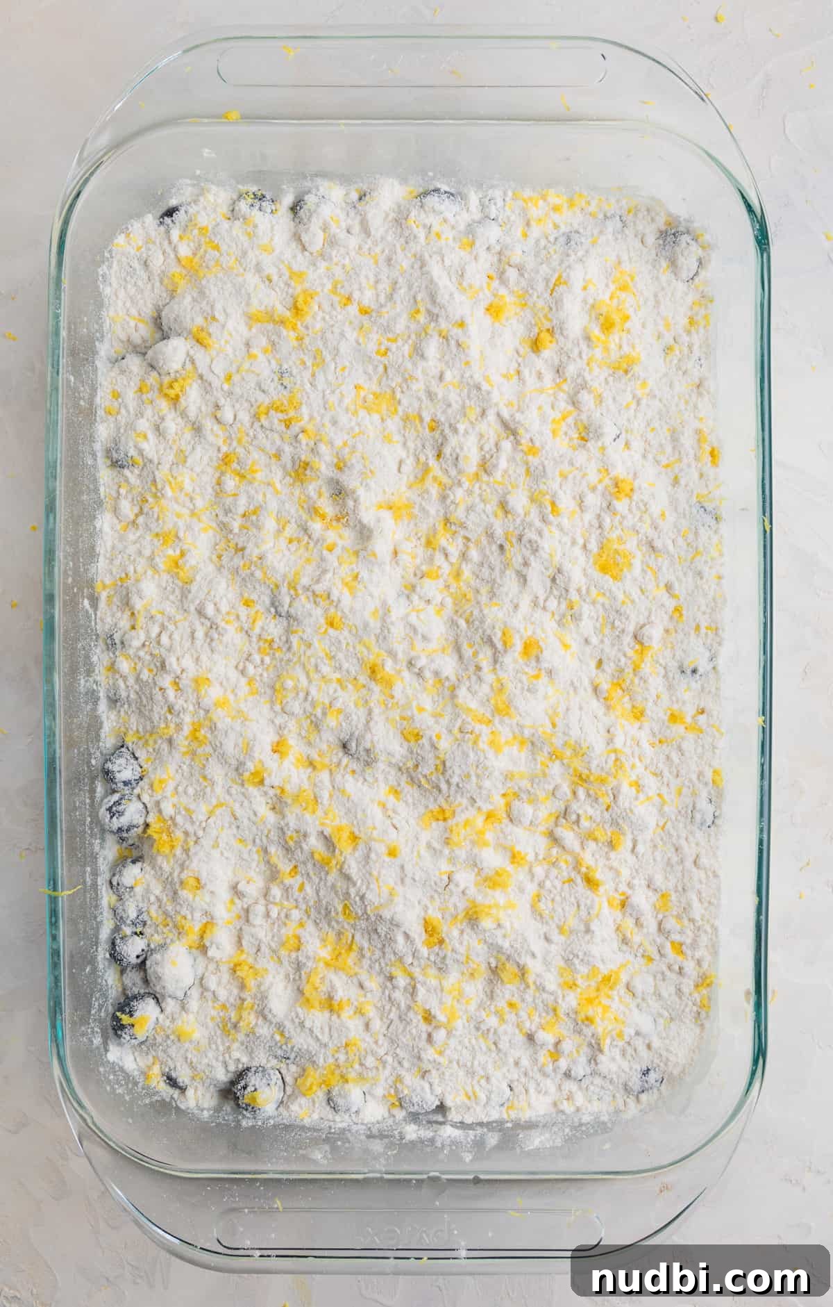 Lemon Blueberry Dump Cake Recipe for Quick Summer Desserts 6 Lemon zest over top of cake mix that is covering blueberries in baking pan.