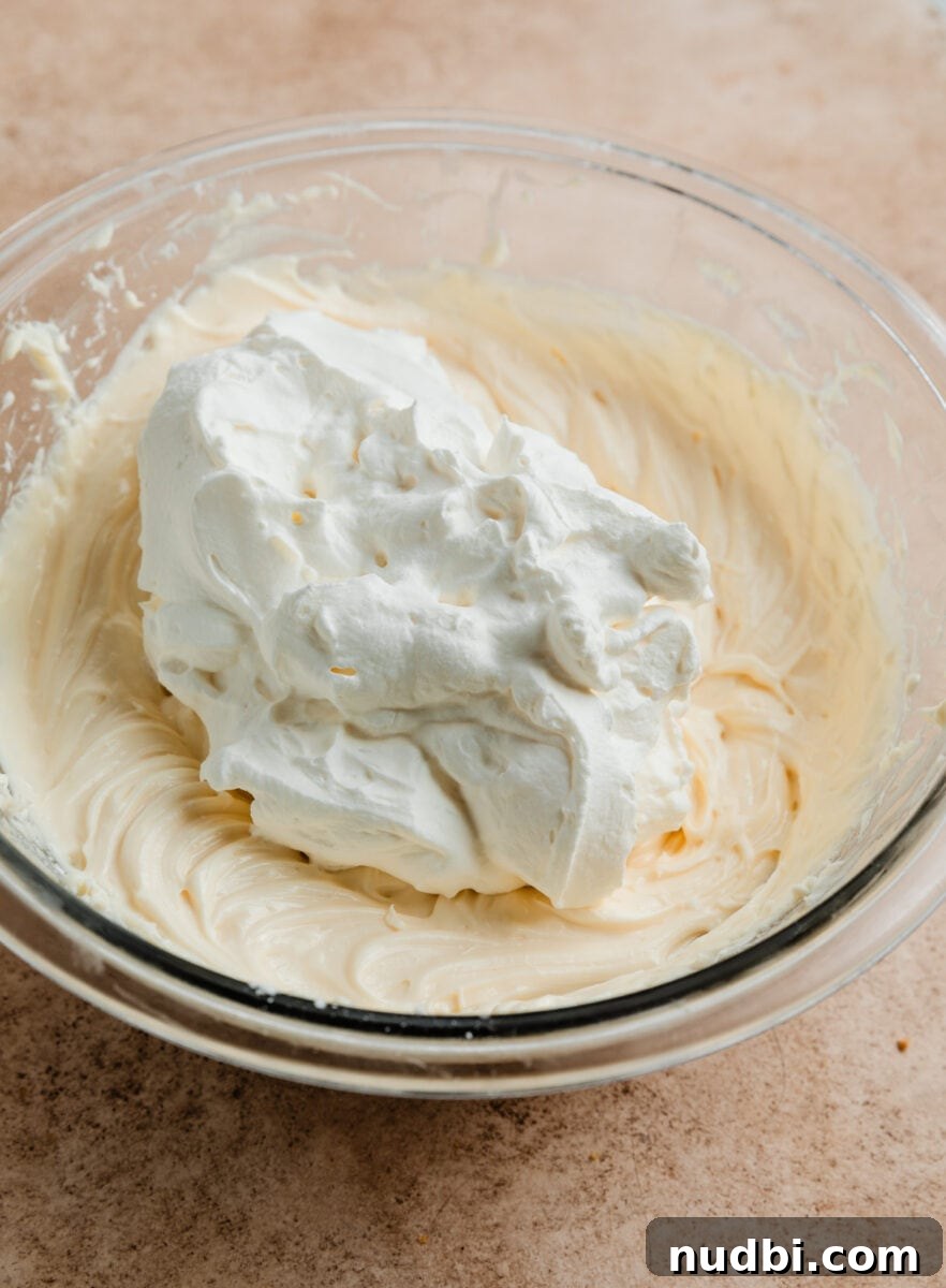 No-Bake Caramel Apple Cheesecake Recipe for Fall Gatherings 12 Whipping cream added to cheesecake mixture in glass bowl.