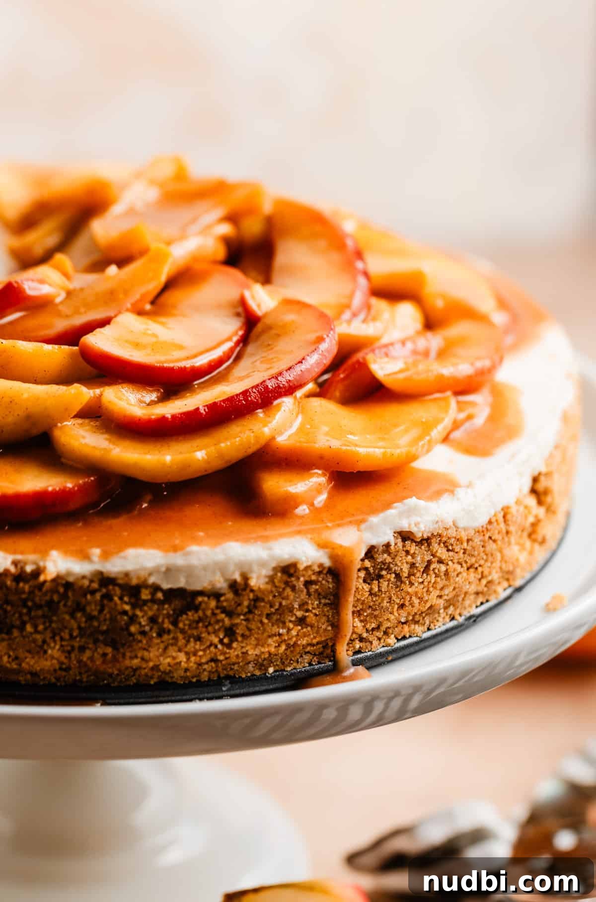 No-Bake Caramel Apple Cheesecake Recipe for Fall Gatherings 18 Caramel apple cheesecake on white cake stand with caramel dripping off the side.