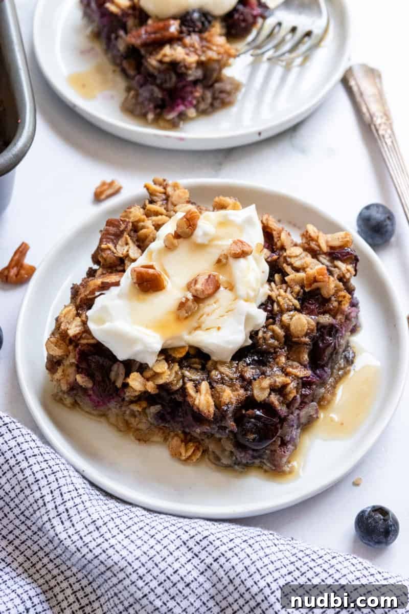 Blueberry Baked Oatmeal Recipe for Cozy Breakfasts 2 Slice of blueberry baked oatmeal on white plate with maple syrup.