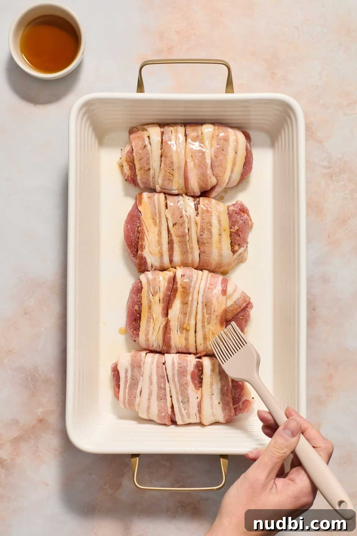 Maple-Glazed Bacon-Wrapped Pork Chops with Crispy Edges 7 Maple being brushed on bacon wrapped pork chops in baking dish with dish of maple syrup beside the pan.