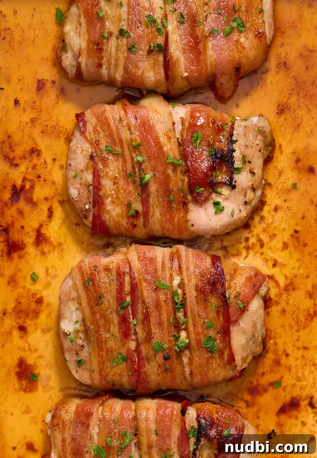 Maple-Glazed Bacon-Wrapped Pork Chops with Crispy Edges 9 Close up view of bacon wrapped pork chops in pan topped with chopped parsley.