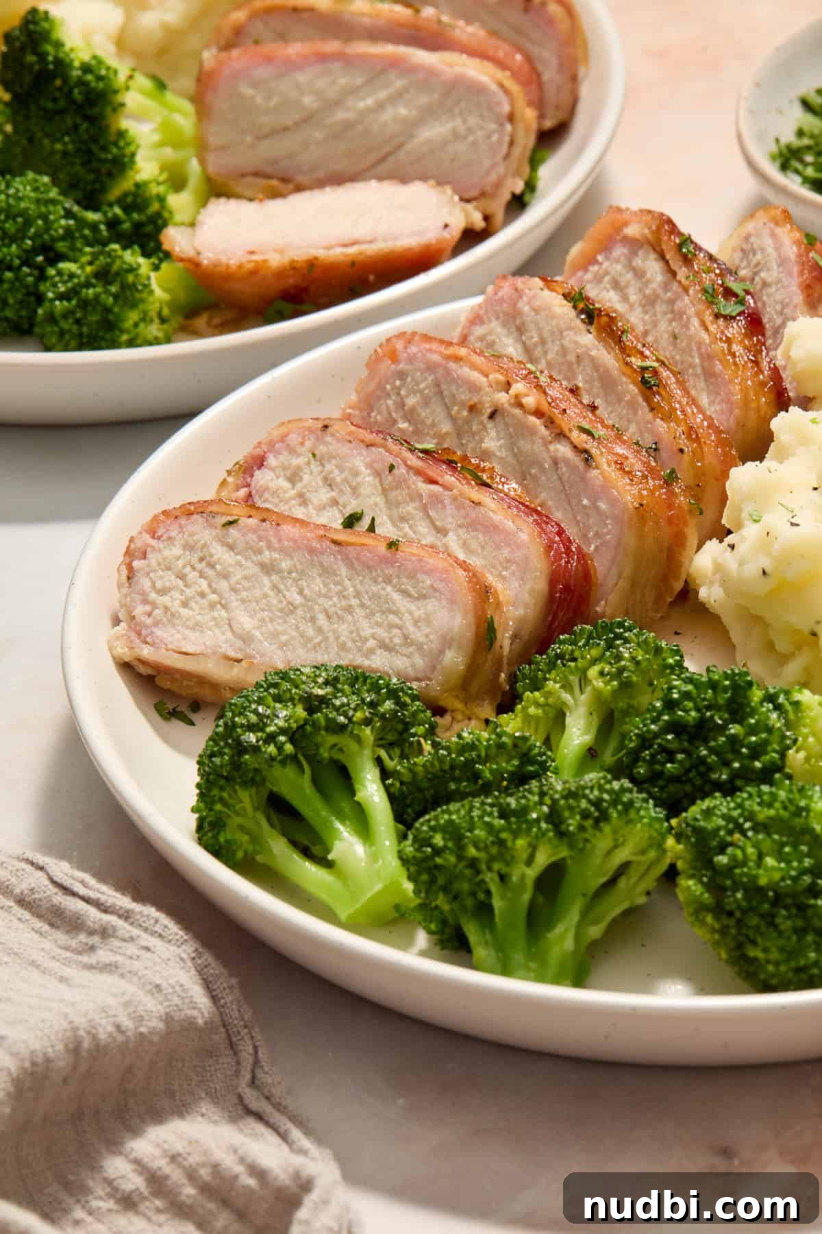 Maple-Glazed Bacon-Wrapped Pork Chops with Crispy Edges 10 Sliced of pork chop wrapped in bacon on dish with broccoli and potatoes.