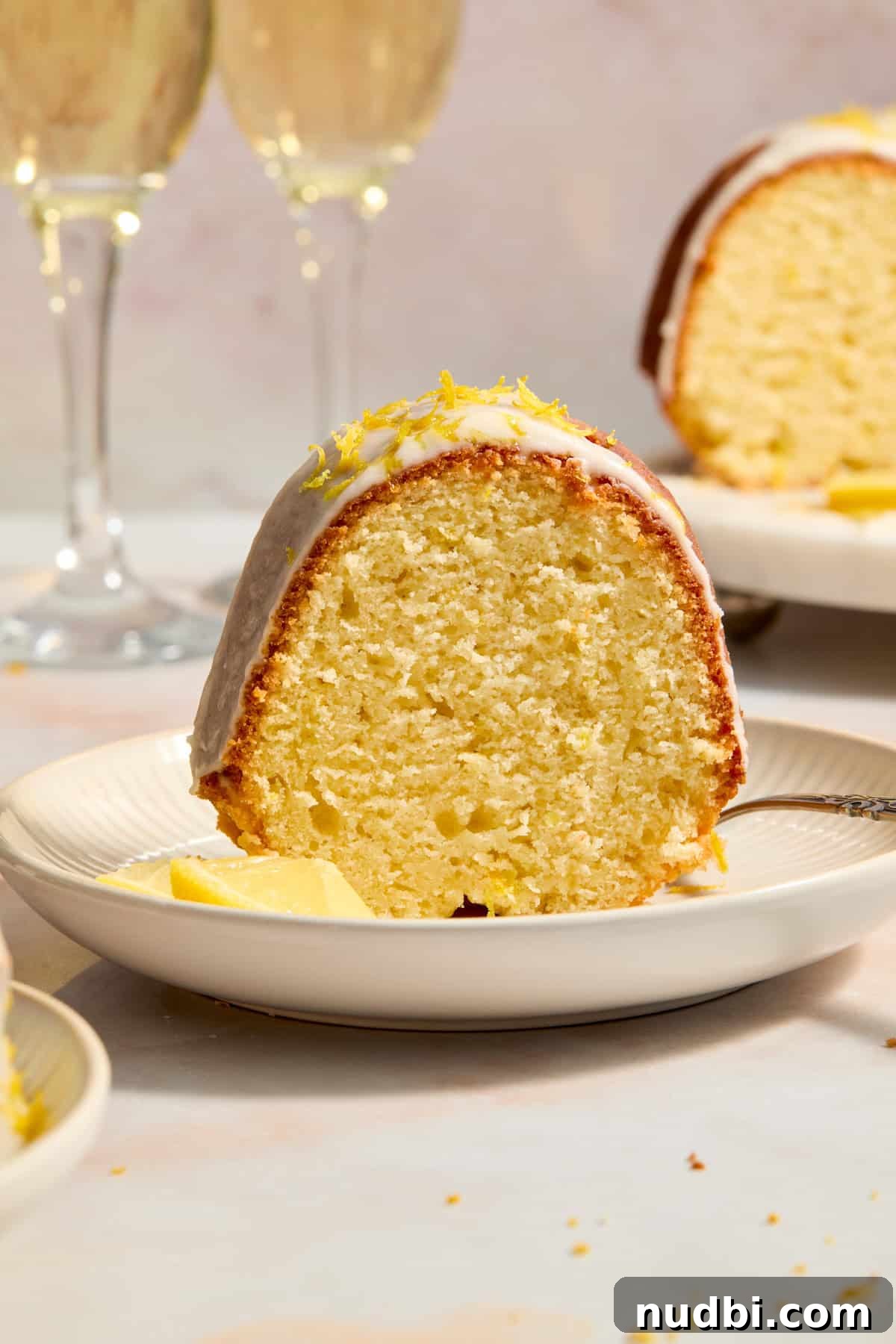 Champagne-Infused Lemon Bundt Cake Recipe 9 Slice of lemon bundt cake on plate with lemon slices and glasses of champagne in the background.