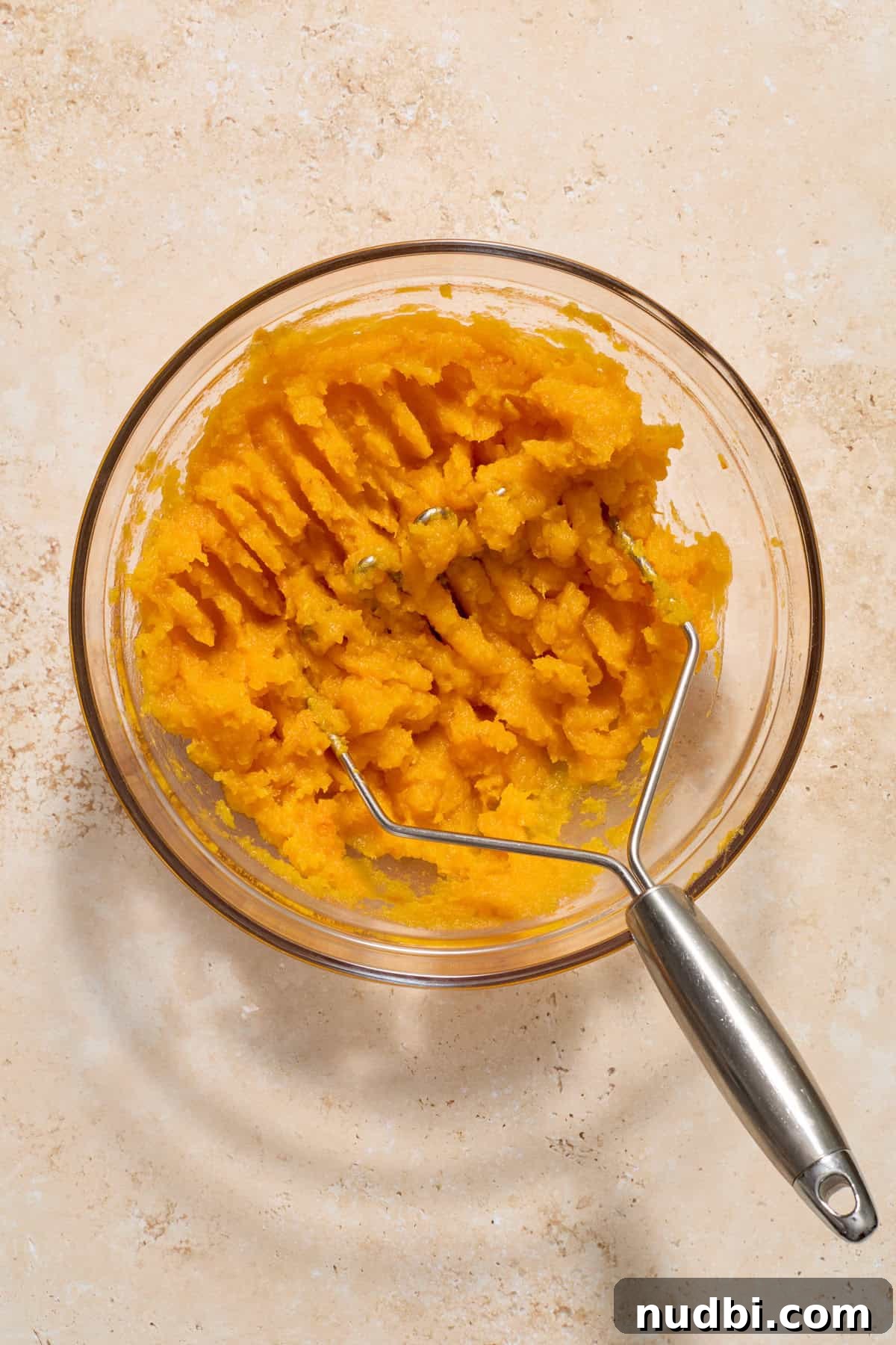 Creamy Butternut Squash Casserole with Sage and Parmesan 5 Mashed butternut squash in glass mixing bowl with potato masher.
