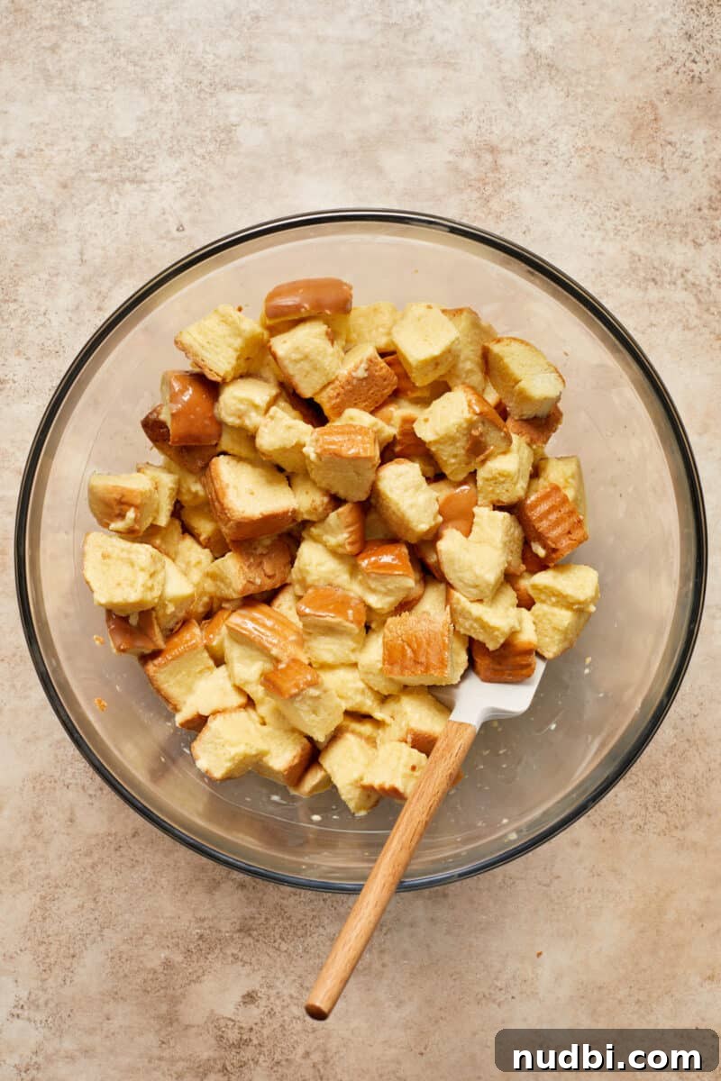 Cinnamon Roll Baked French Toast Recipe for Brunch 6 Egg mixture combined with cubed Brioche bread with spatula in glass mixing bowl.