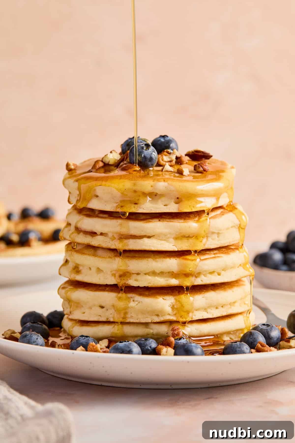 Stack of thick fluffy pancakes on plate with maple syrup drizzling over top of pancakes.