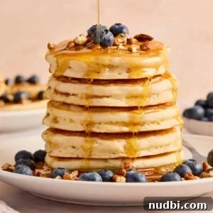 Stack of super fluffy pancakes on plate with blueberries, pecans, and maple syrup.