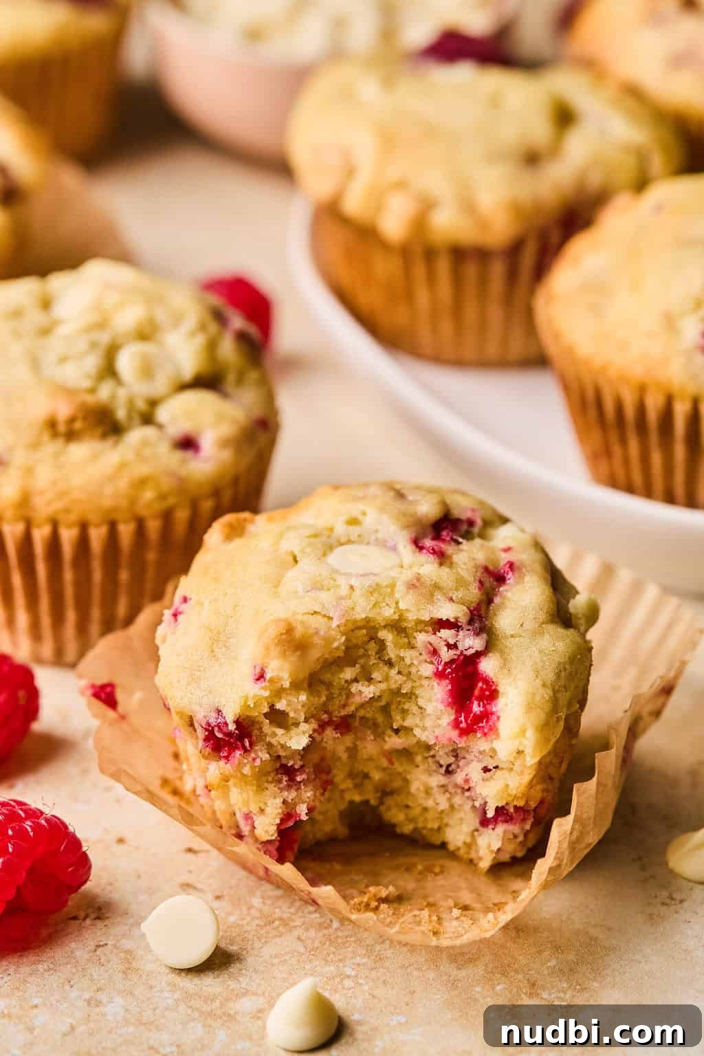 White Chocolate Raspberry Muffins Recipe — Moist, Fruity Treat 2 White chocolate raspberry muffins on surface with bite taken out of front muffin.