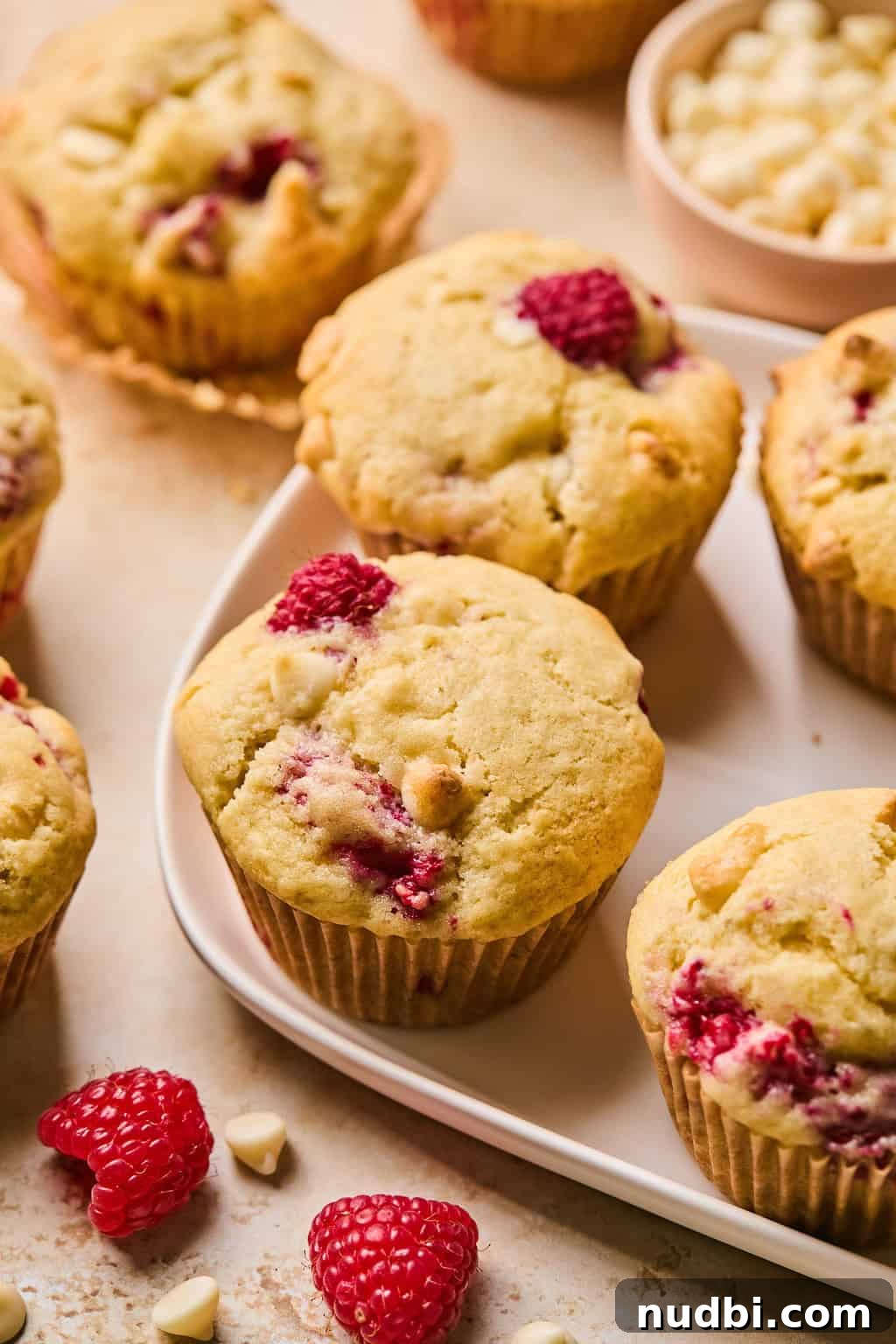 White Chocolate Raspberry Muffins Recipe — Moist, Fruity Treat 12 Raspberry muffins with white chocolate chips on plate with white chocolate chips and raspberries surrounding the plate.