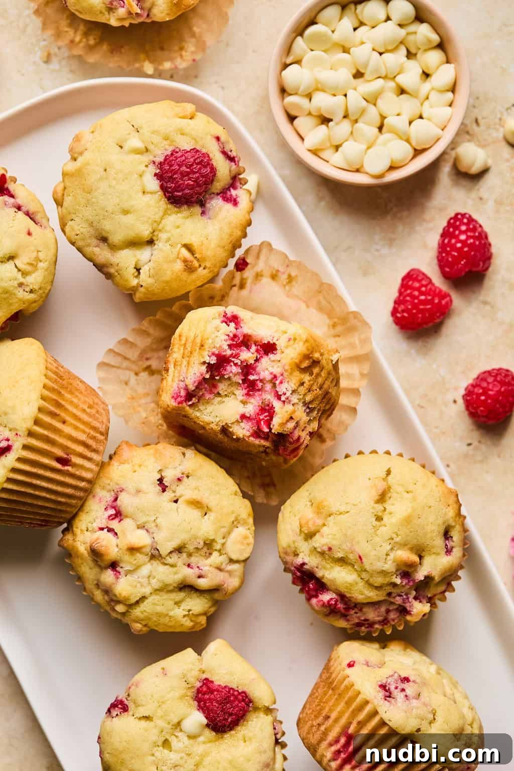 White Chocolate Raspberry Muffins Recipe — Moist, Fruity Treat 4 White plate with raspberry and white chocolate chip muffins with bite taken out of center muffin.