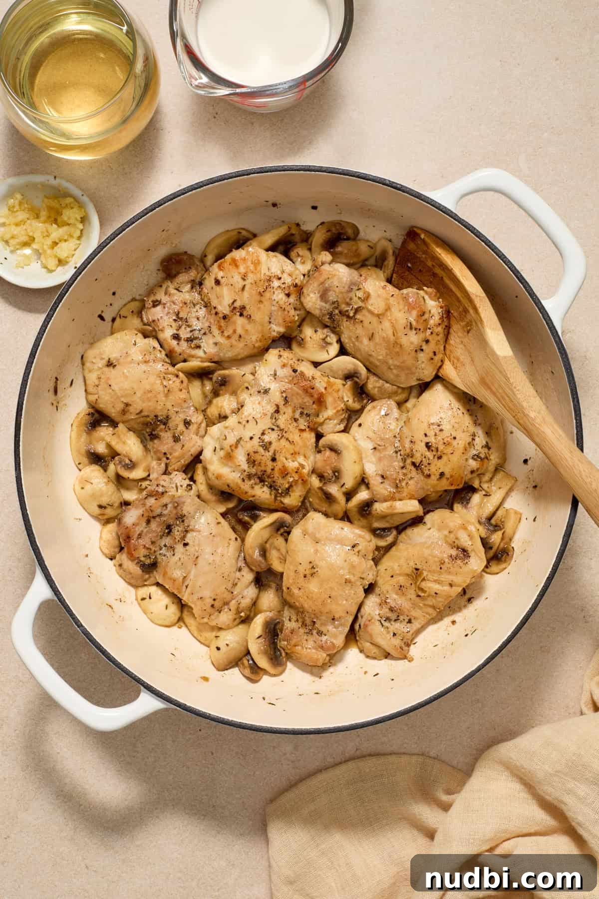 Creamy Mushroom Chicken Thighs with Garlic and Herbs 6 Mushrooms added to skillet with chicken thighs and wooden spoon propped in the pan.