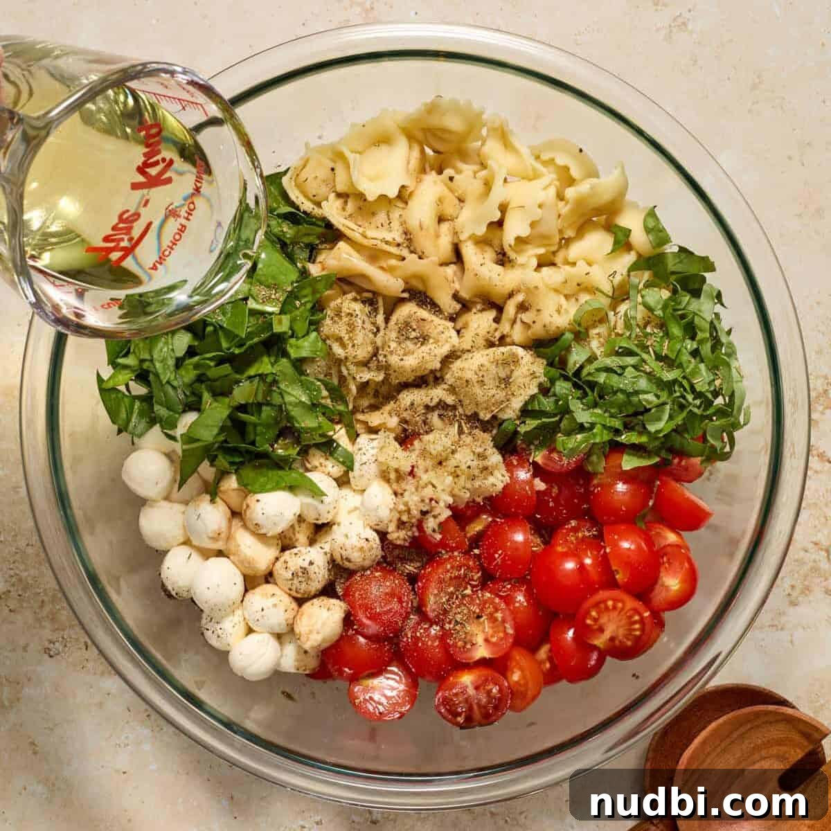 Caprese Tortellini Salad with Cherry Tomatoes and Basil 7 Pasta salad ingredients in bowl with oil being poured over.