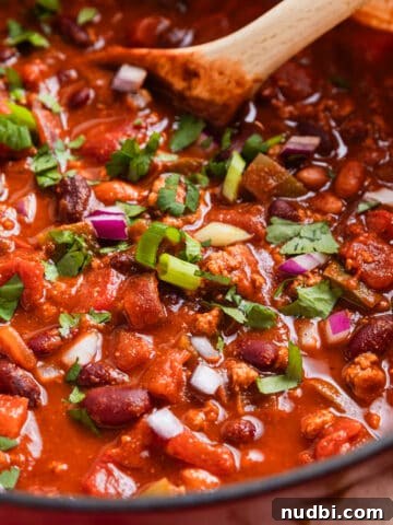 Red dutch oven with ground turkey chili topped with red onion, green onion and cilantro with wooden spoon sitting in pot.