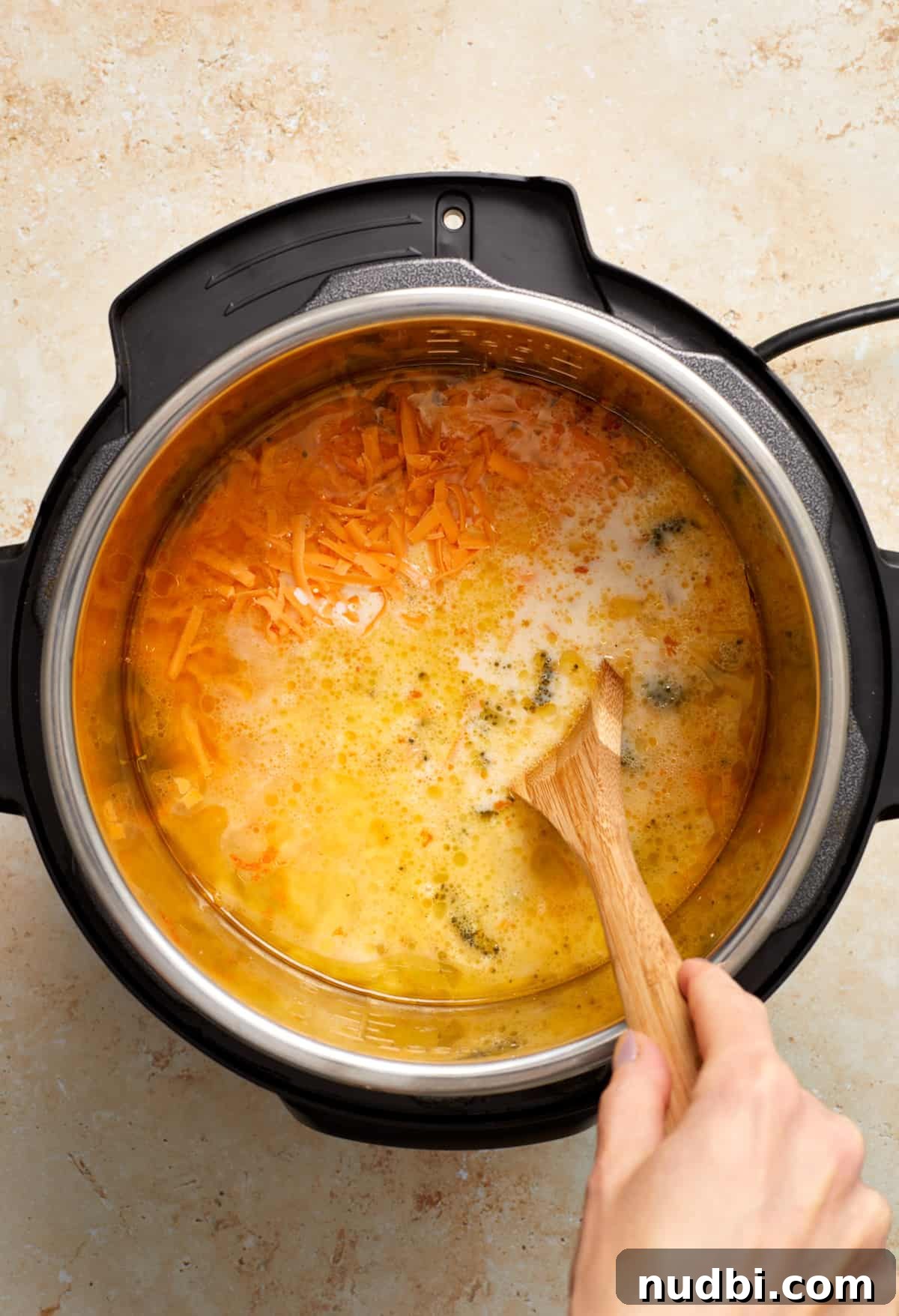 Creamy Broccoli Cheddar Soup Made in the Instant Pot 7 Cheese and cream added to soup pot with other ingredients and being stirred with wooden spoon.