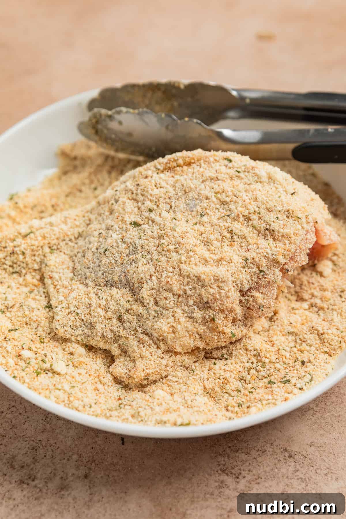 Crispy Parmesan-Crusted Air Fryer Chicken Thighs 5 Chicken thigh with tongs in bread crumb and parmesan mixture.