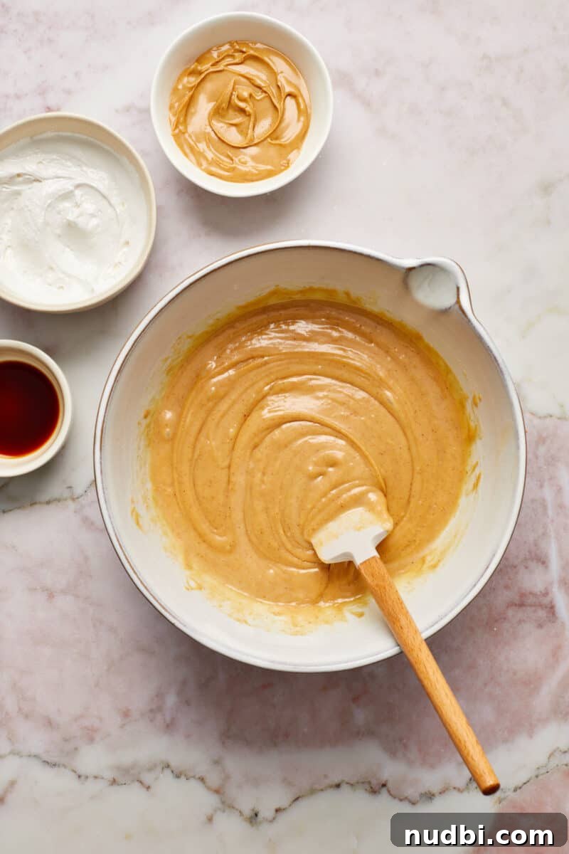 Quick Microwave Peanut Butter Fudge Recipe 5 Peanut butter mixture in bowl with spatula with bowls of marshmallow cream, peanut butter and vanilla beside it.