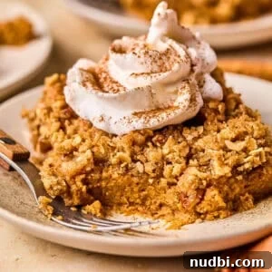Slice of pumpkin pie crumble on plate with whipped cream and cinnamon over top and a fork on the plate beside it.
