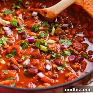 Cozy Dinner Recipes for Comforting Weeknight Meals 25 Red dutch oven with ground turkey chili topped with red onion, green onion and cilantro with wooden spoon sitting in pot.