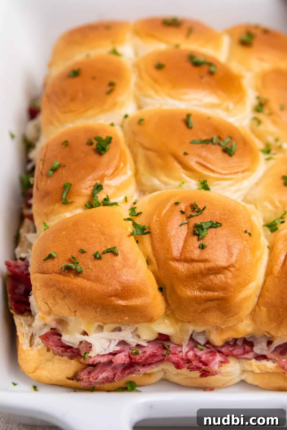 Classic Reuben Sliders with Melted Swiss and Tangy Sauerkraut 2 Slow cooker reuben sliders in white pan with chopped parsley on top.