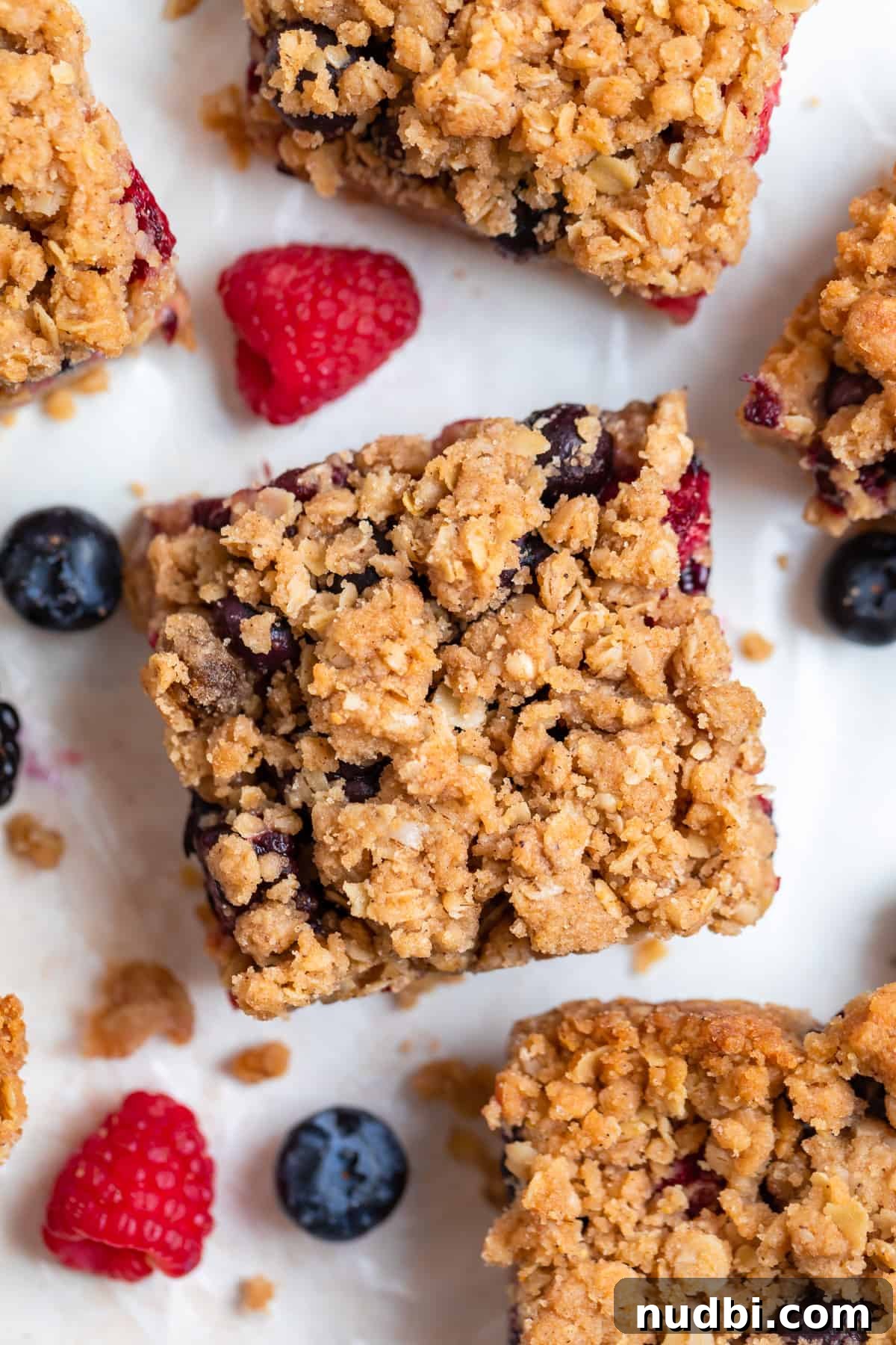 Mixed Berry Crumble Bars Recipe with Oat Streusel Topping 2 Berry crumble bars with oat topping on parchment with fresh berries surrounding.