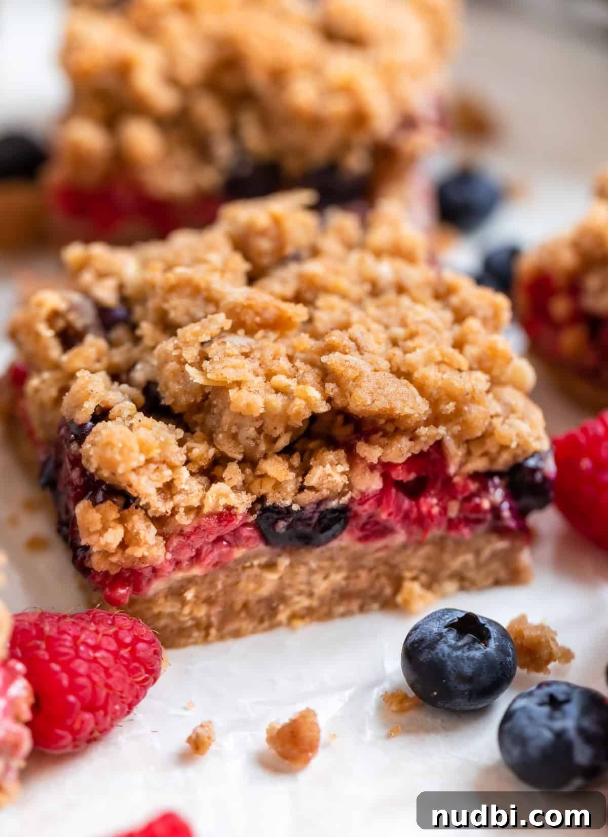 Mixed Berry Crumble Bars Recipe with Oat Streusel Topping 3 Mixed berry bar on wax paper with blueberries and raspberries surrounding.