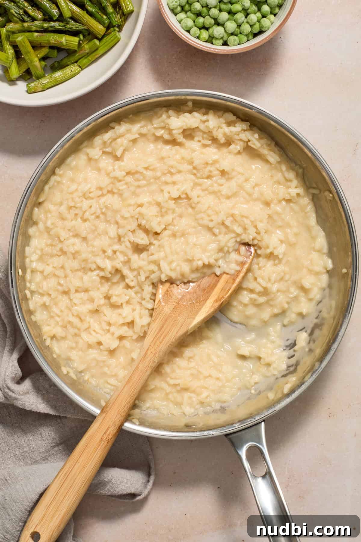 Creamy Spring Risotto with Peas and Asparagus 11 Creamy risotto with parmesan cheese in skillet with wood spoon and peas and asparagus prepped and ready to be added in.