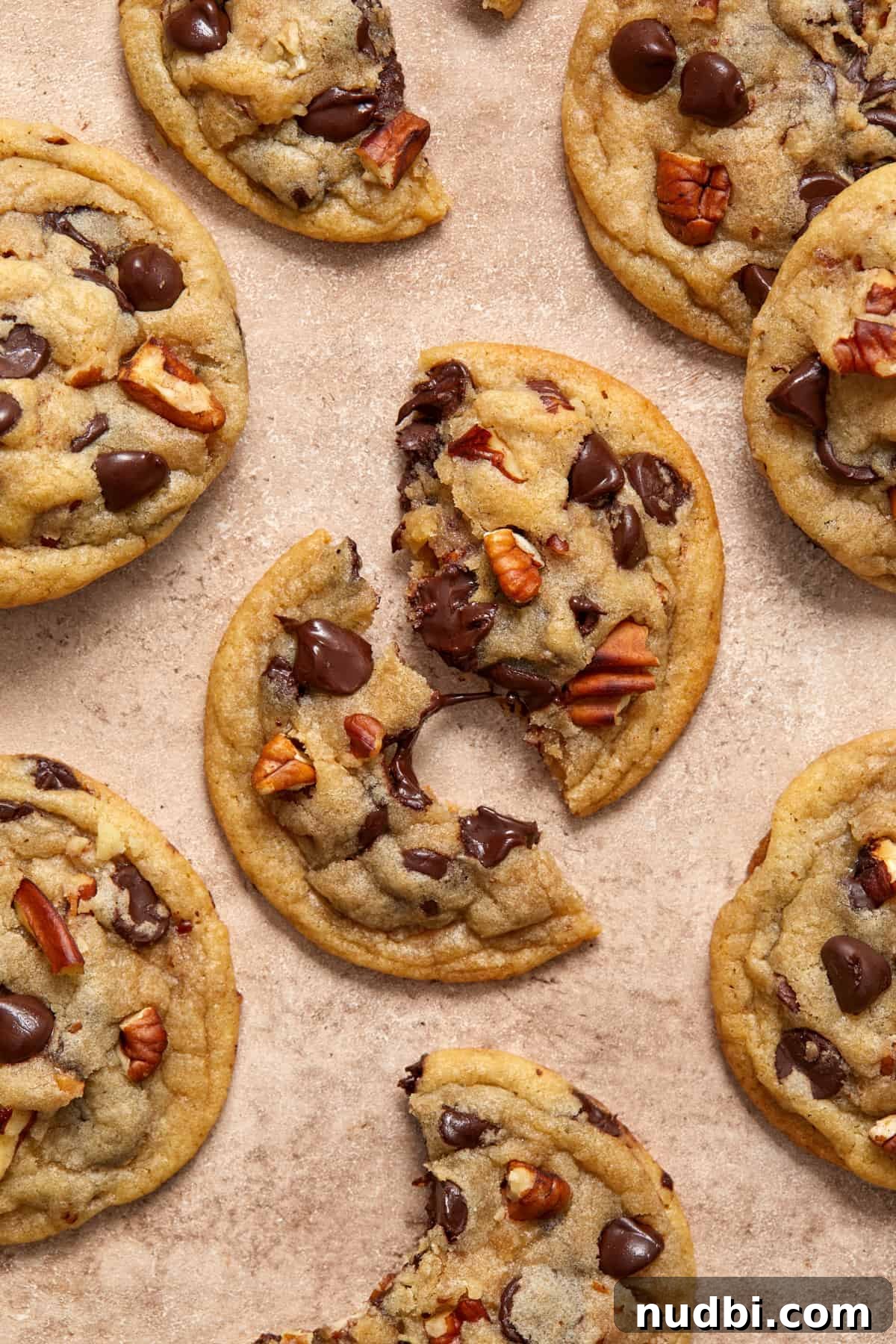 Chewy Chocolate Chip Pecan Cookies Recipe 8 Chocolate chip cookies with pecans on surface with focus on center cookie broken in half with melted chocolate string connecting the pieces.