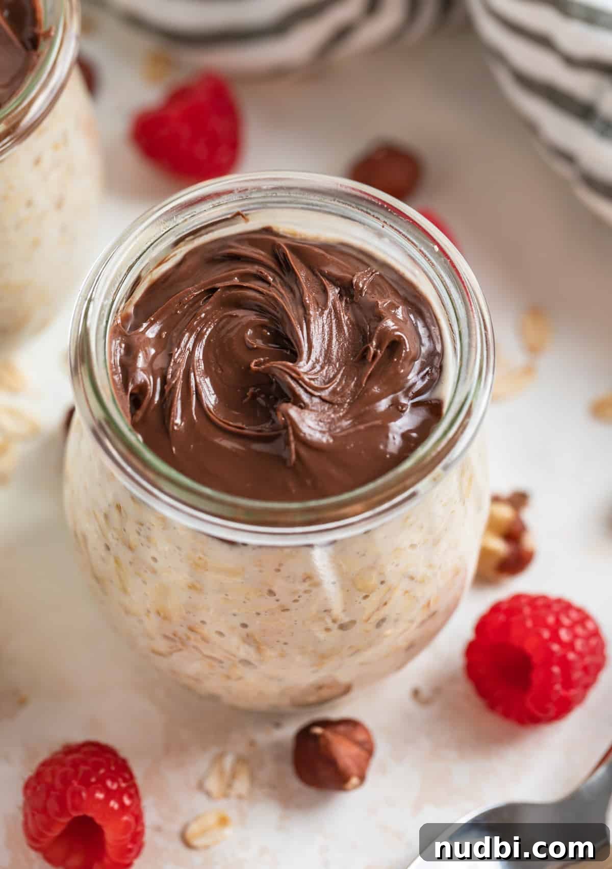 Creamy Nutella Overnight Oats Recipe for Quick Morning Meals 2 Jar of overnight oatmeal with nutella swirled on top and raspberries and hazelnuts beside.