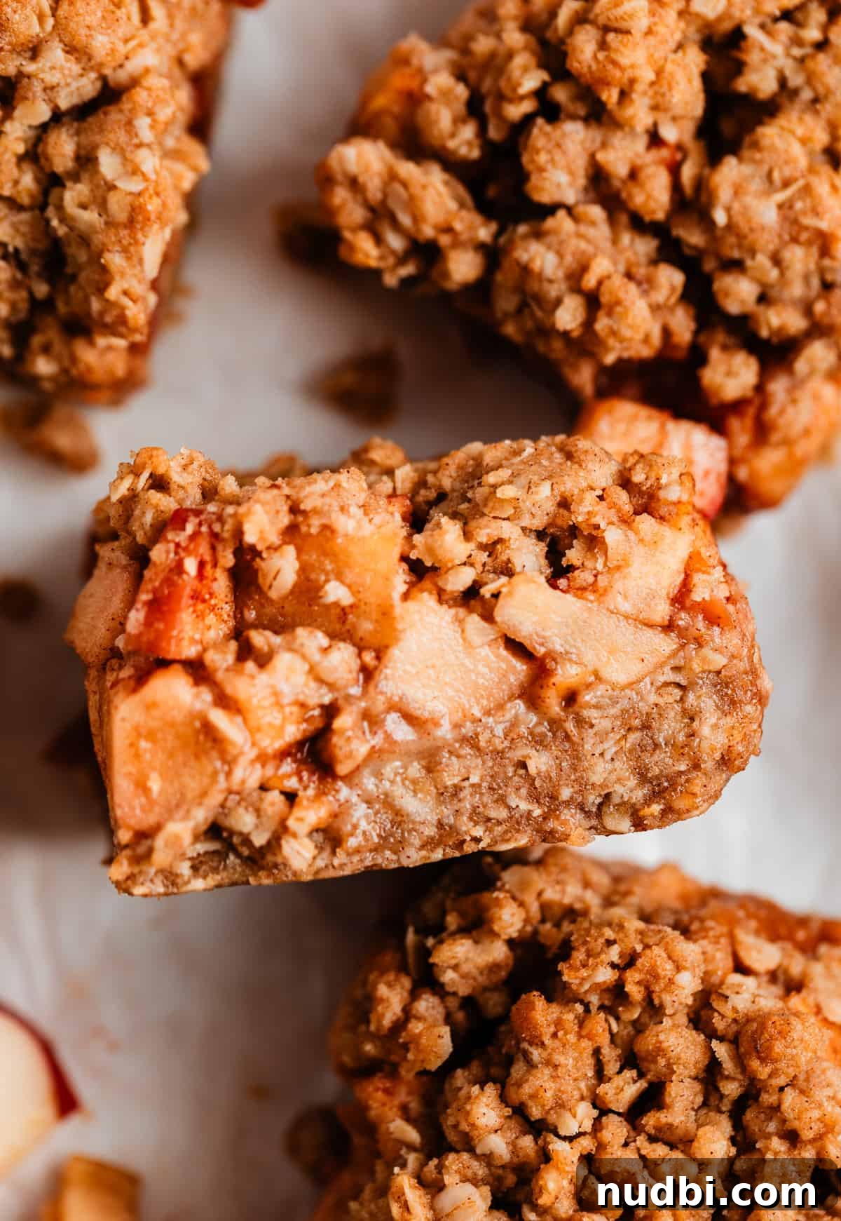 Homemade Apple Crumble Bars Recipe — Soft Filling & Crunchy Topping 2 Apple crumble bars on parchment with layers of apple and oat crumb showing.
