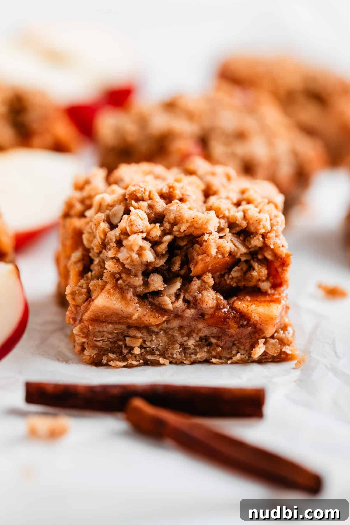 Homemade Apple Crumble Bars Recipe — Soft Filling & Crunchy Topping 8 Layered apple crumble bar slice on parchment with cinnamon sticks.