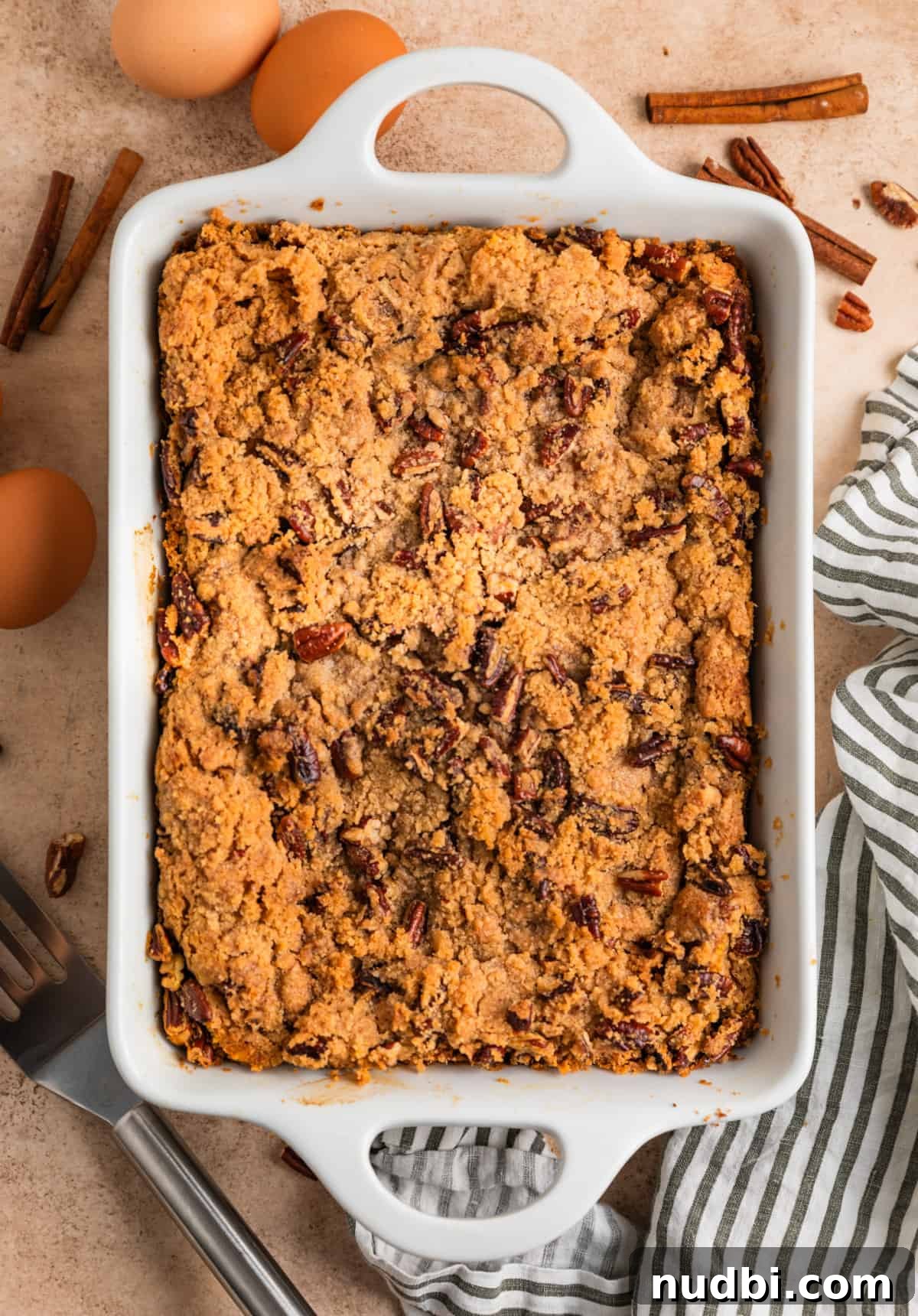 Pumpkin Spice French Toast Casserole Recipe for Fall Brunch 10 White pan with streusel topped baked French toast with striped linen, eggs and spatula beside the dish.