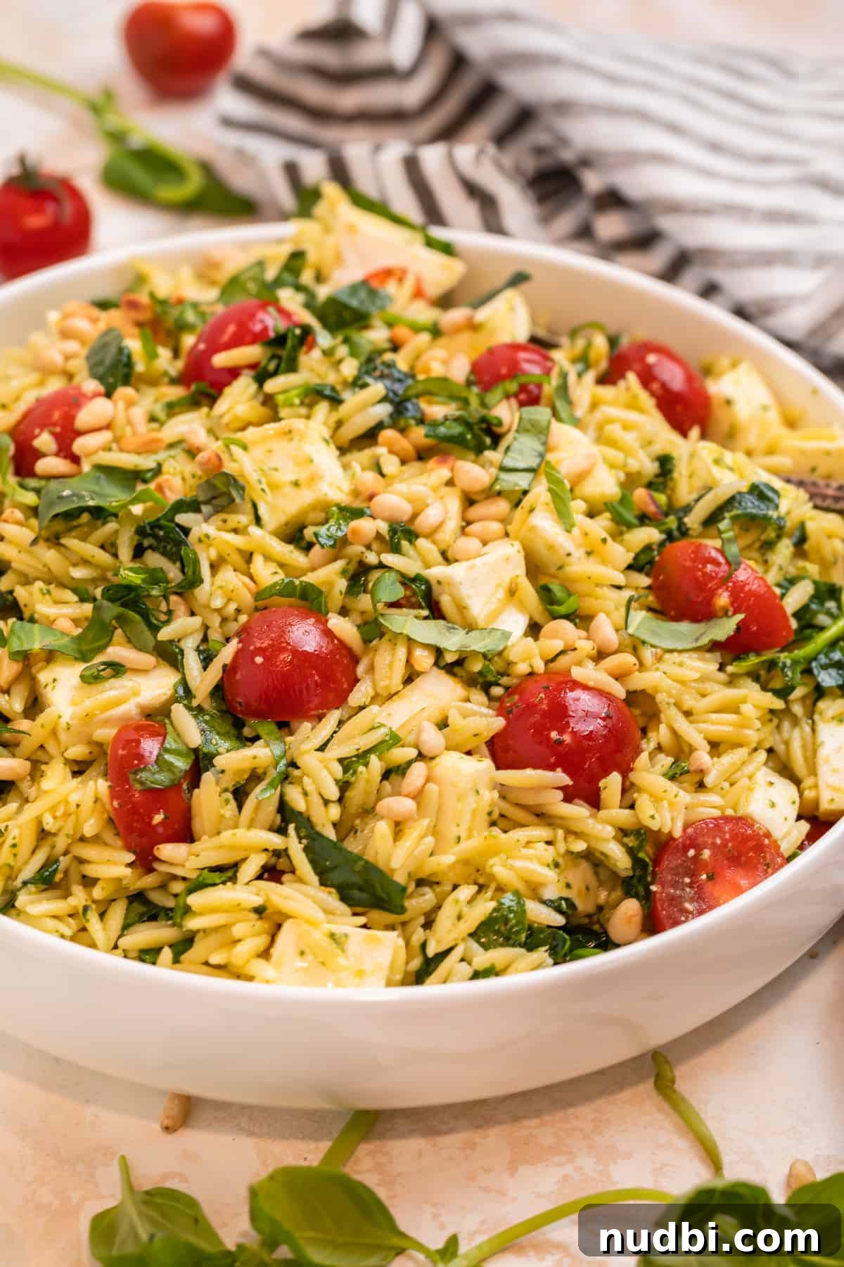 Creamy Pesto Orzo Salad with Cherry Tomatoes and Basil 3 Orzo salad with pesto, mozzarella, tomatoes and chopped spinach in white bowl with towel beside it.