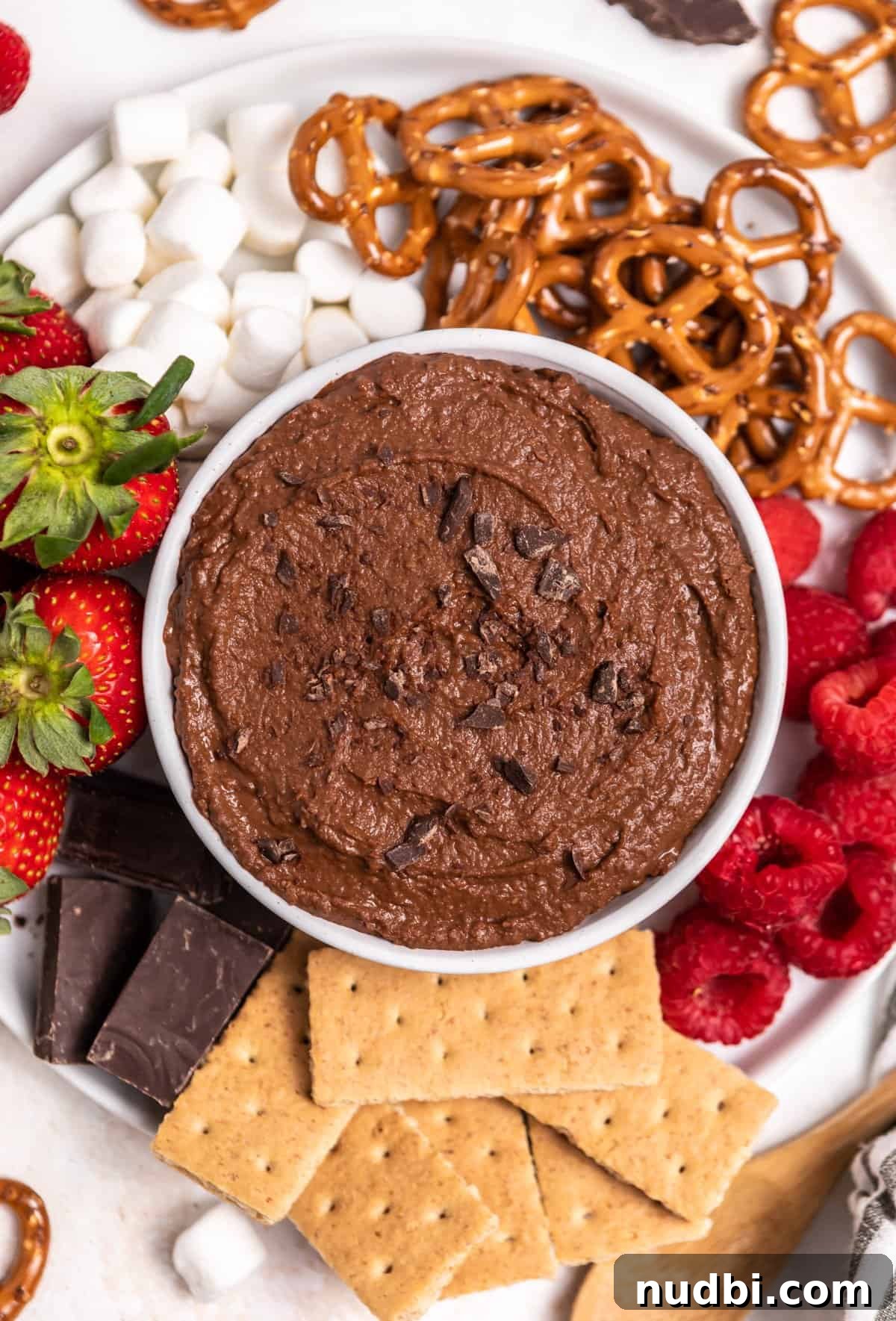 Decadent Brownie Batter Hummus Recipe: Dessert Dip Made Vegan 2 Bowl of chocolate brownie batter hummus with chopped chocolate on top on plate with marshmallows, pretzels and berries.