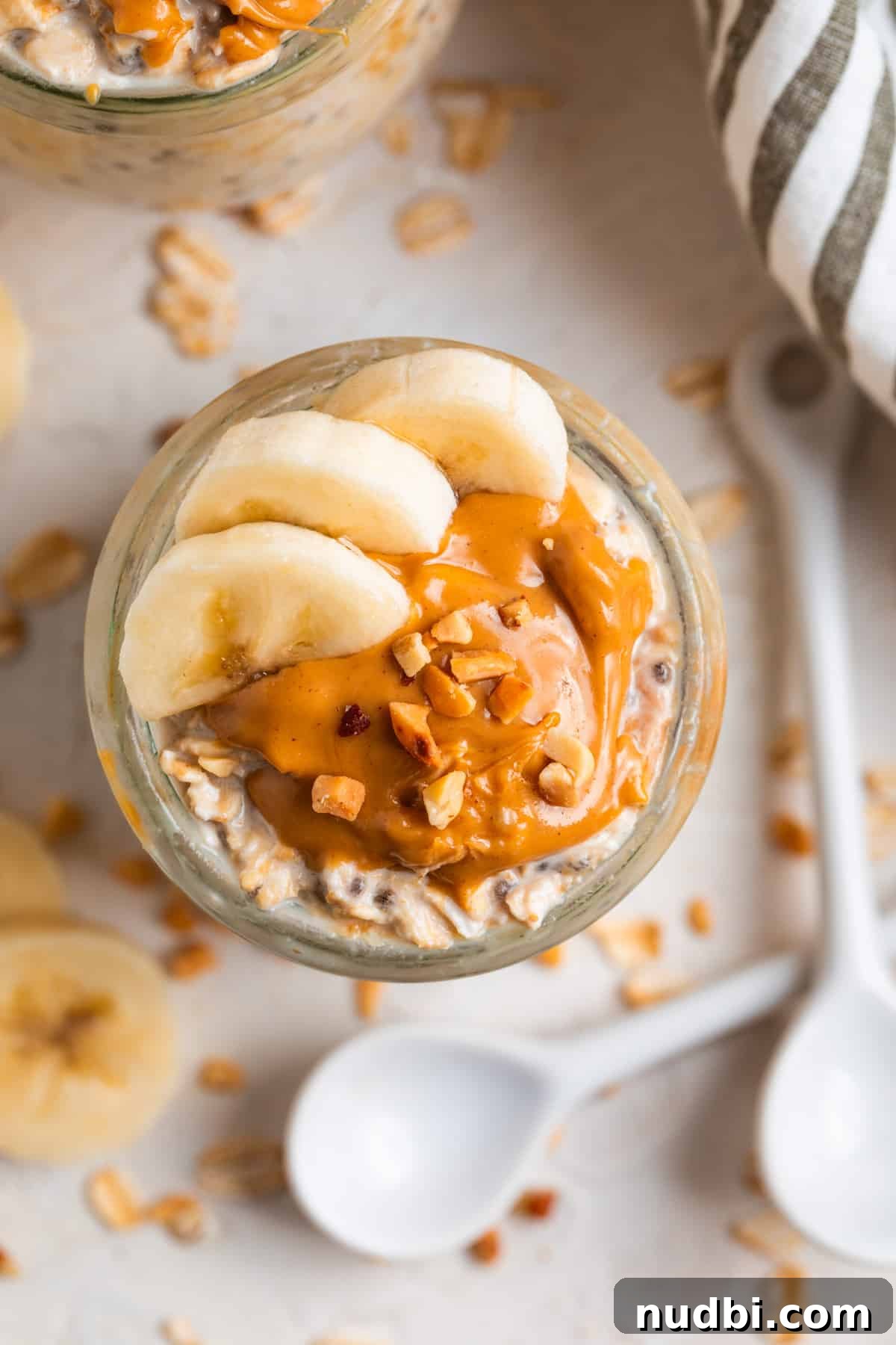 High-Protein Overnight Oats Recipe for Busy Mornings 3 Overhead view of oatmeal topped with sliced bananas and peanut butter with white spoon and rolled oats surrounding.